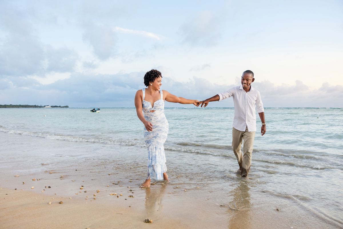 A couple in white attire walks playfully along a sandy beach at sunset holding hands and enjoying a romantic moment.