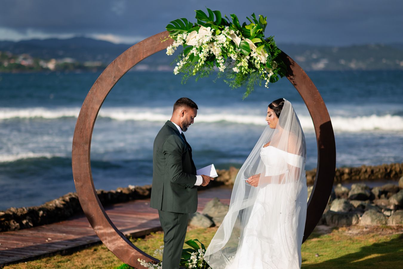Intimate wedding ceremony moment under circular arch with ocean waves in background.