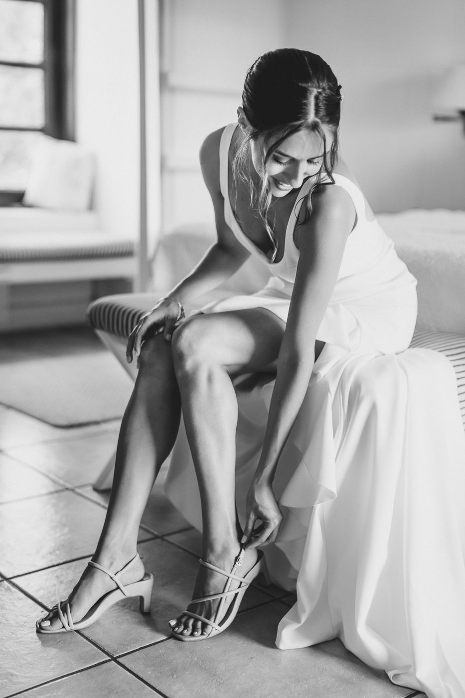 Black and white photo of someone in white dress fastening shoes while sitting.