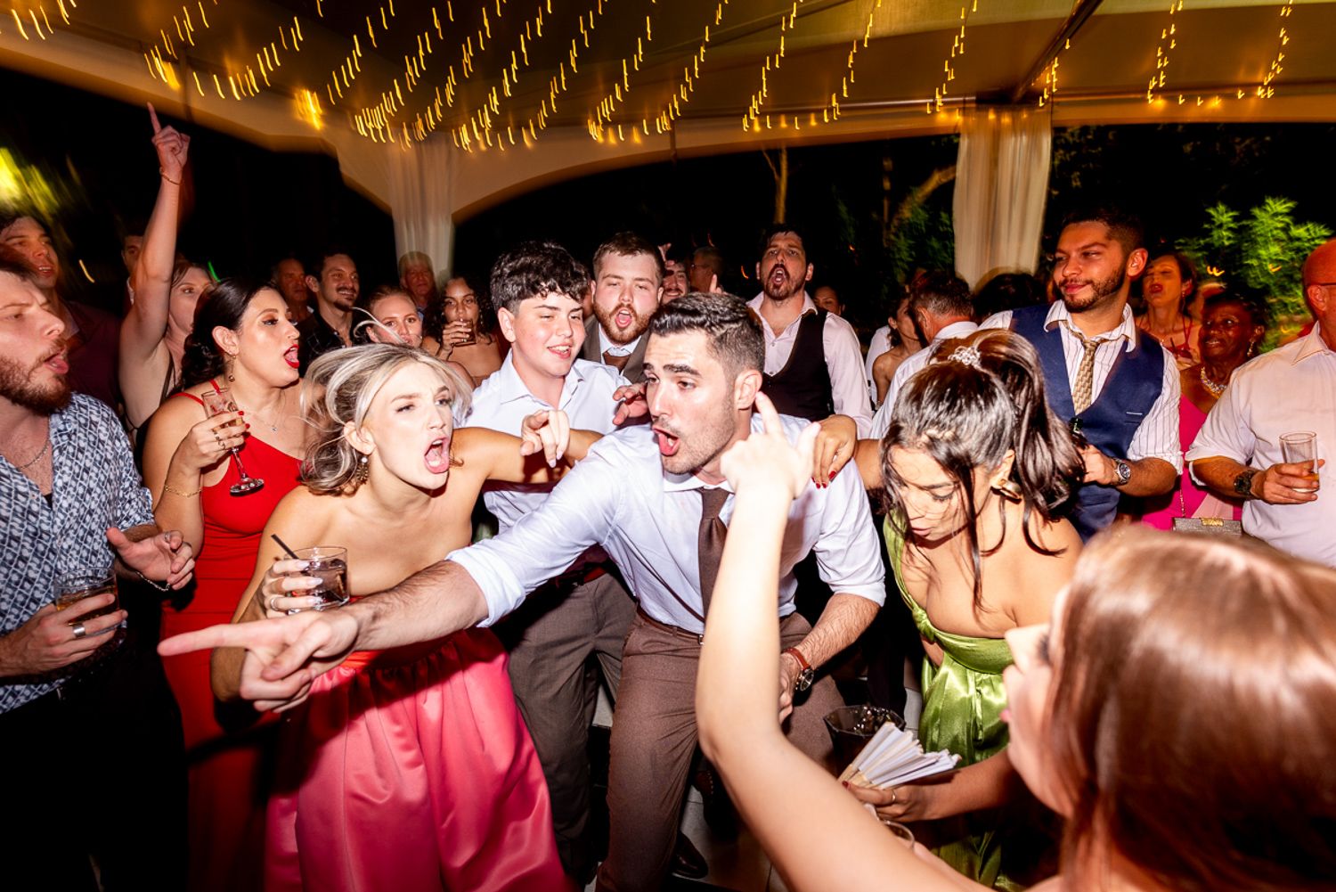 Lively crowd dancing and celebrating together at an indoor wedding reception under warm ambient lighting.