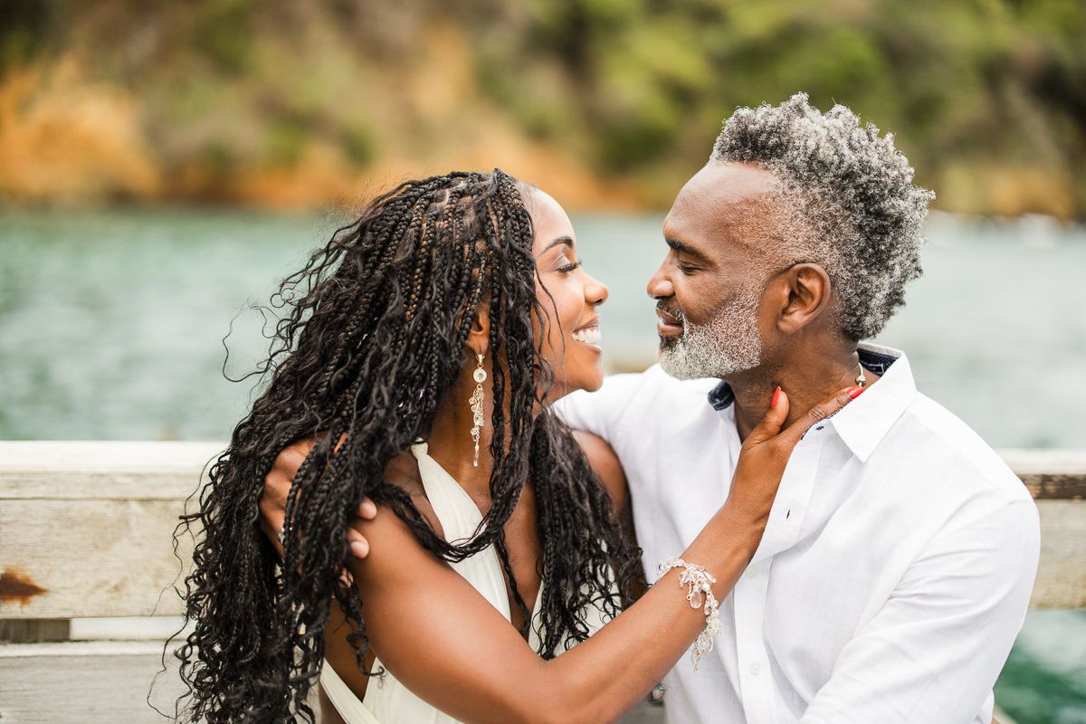 A romantic Black couple shares an intimate moment outdoors near water, the woman with long locs and the man with gray hair and beard.