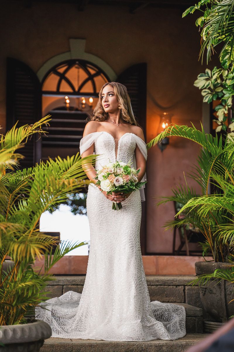 Bride in white off-shoulder lace gown holding floral bouquet stands on steps surrounded by tropical plants at elegant venue.