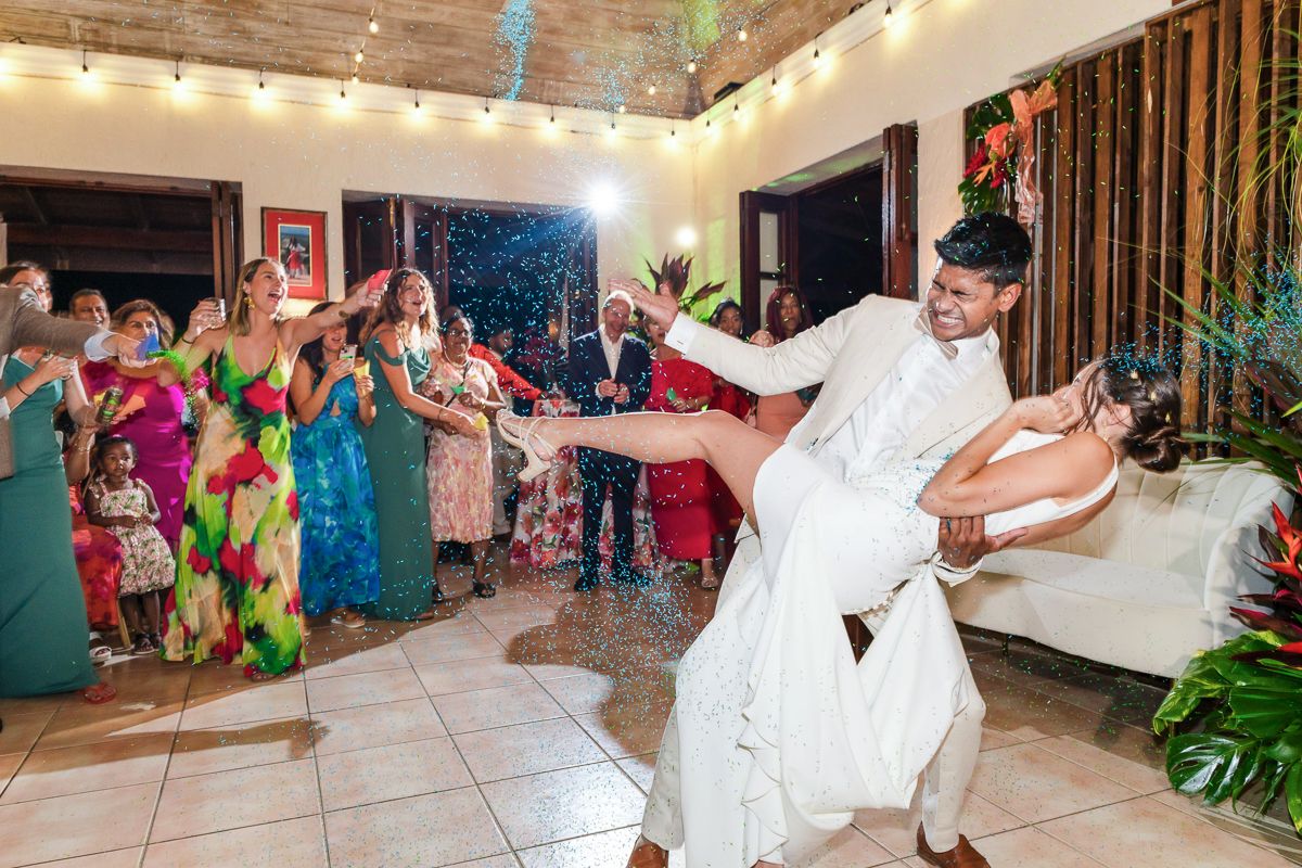 Groom dips bride on dance floor at colorful wedding reception with guests celebrating in background.