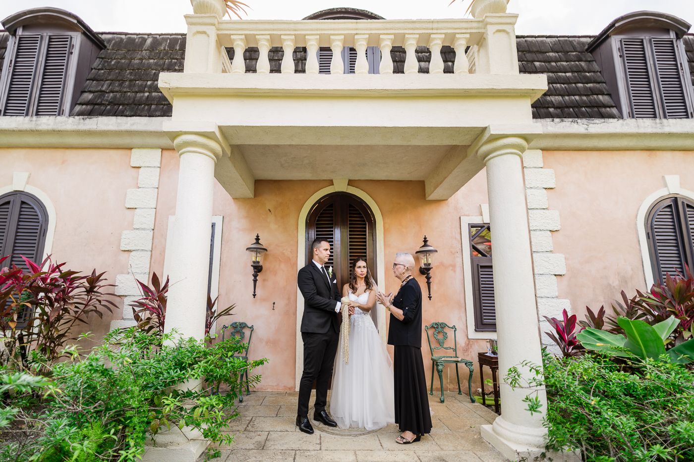 Bride and groom stand with officiant before a pink Mediterranean-style villa during an elegant outdoor wedding ceremony.