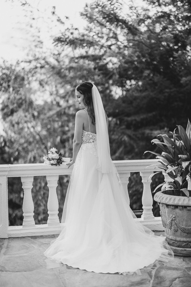Bride in strapless gown with long veil holding bouquet on elegant balcony surrounded by lush greenery.