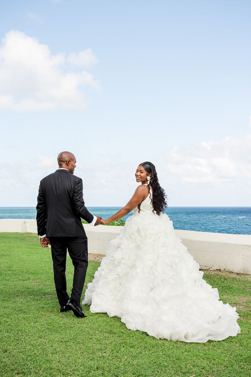 A couple in wedding attire walks together on a grassy oceanfront lawn during their beach wedding celebration.