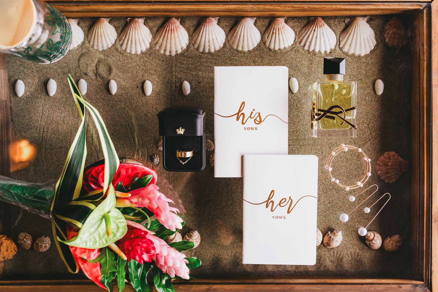 Wedding details flatlay with invitations, seashells, rings, and pink flowers arranged on dark wooden tray.