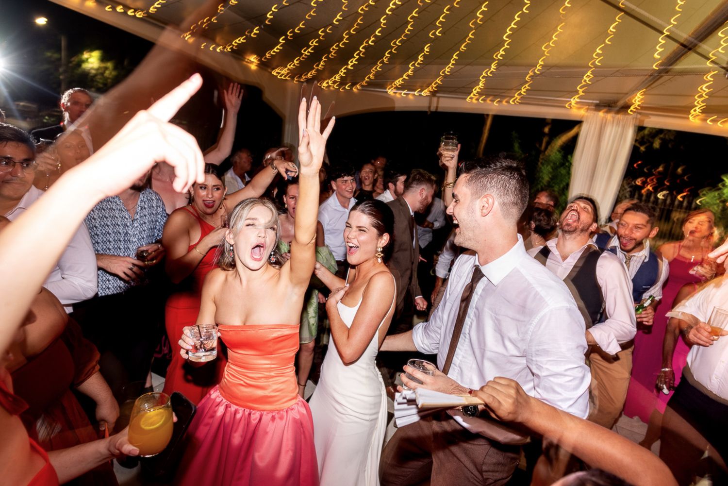 Wedding guests dance and celebrate on a crowded dance floor under twinkling string lights during a lively reception.