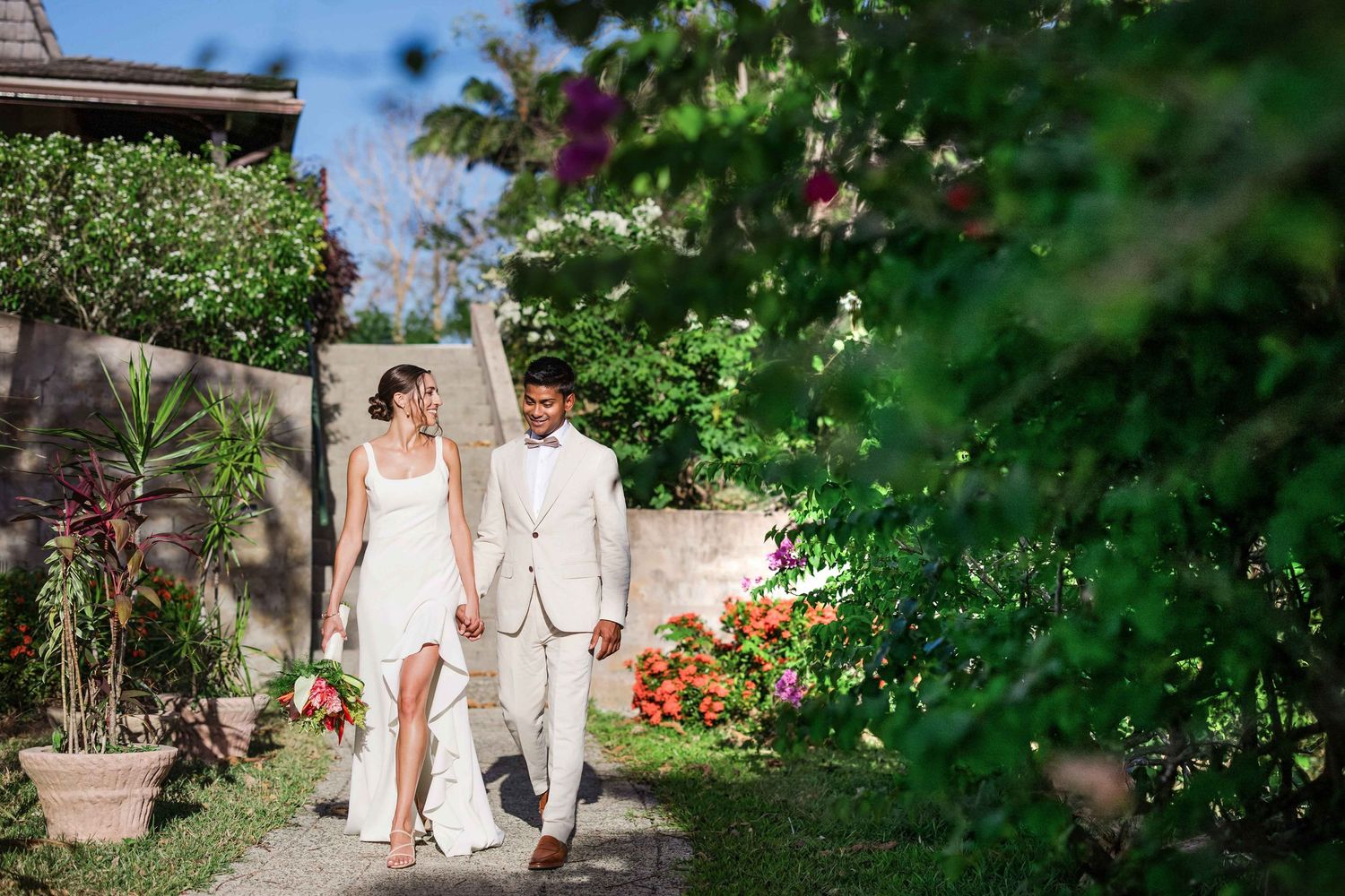 Couple walking through lush garden pathway in wedding attire on their special day.