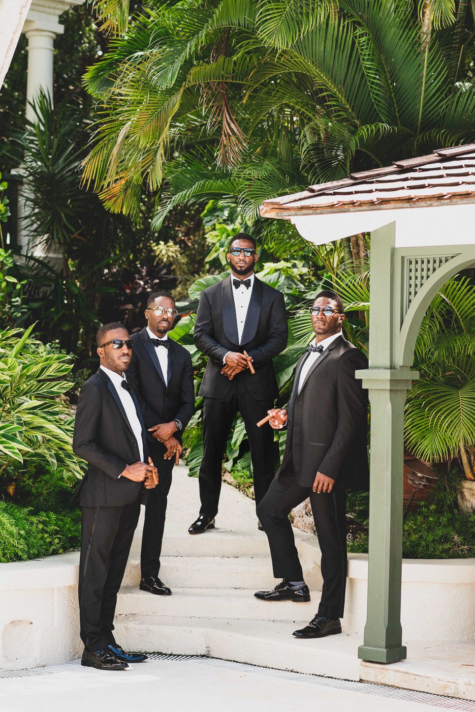 A group in formal black tuxedos posing under a white gazebo surrounded by tropical palm trees and gardens.