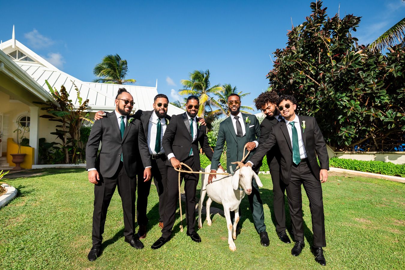 Wedding party jumps joyfully on grass with a white chapel building in the background.
