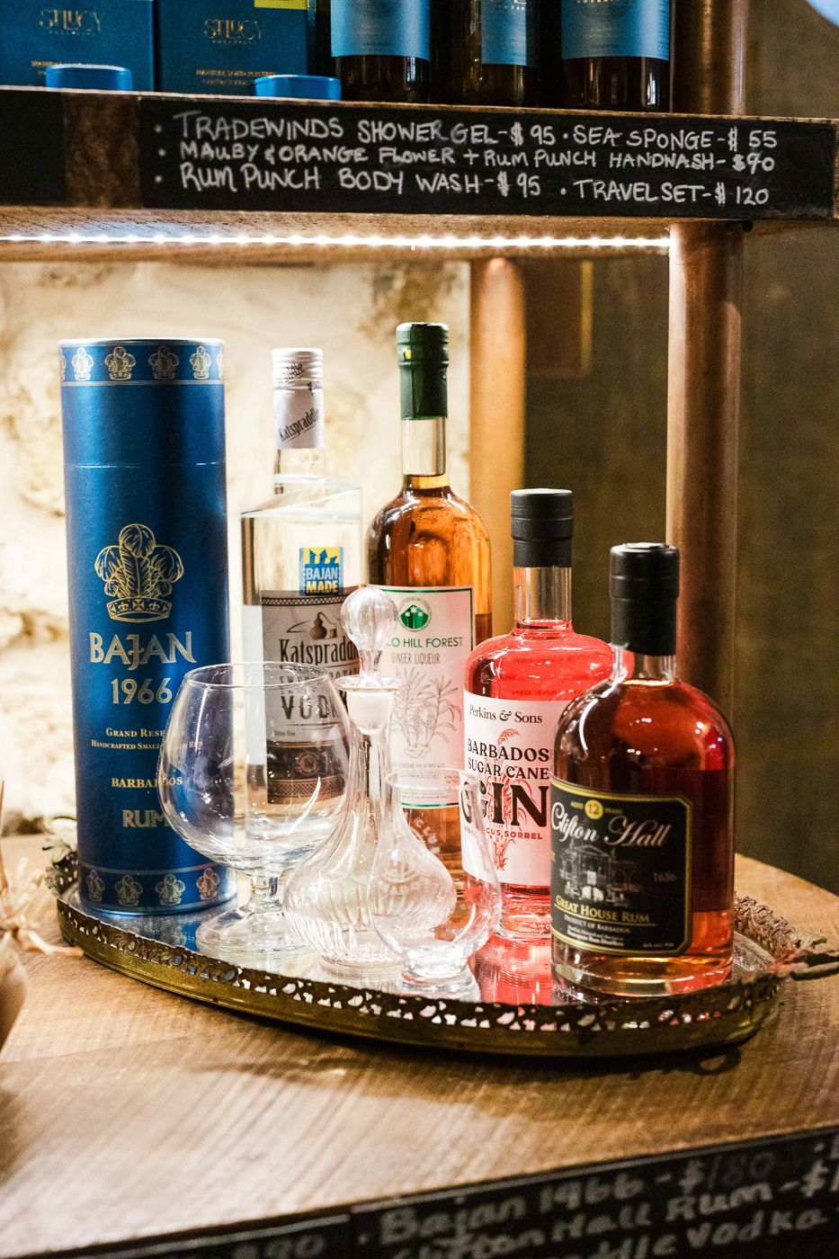 A collection of premium spirits and glasses displayed on a wooden bar tray with menu pricing above.