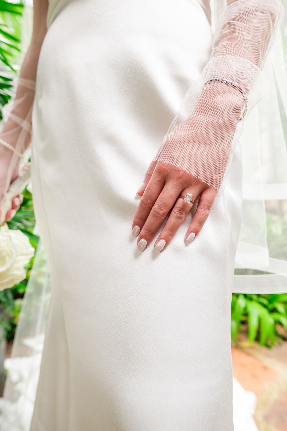 Close-up of bride's hand showing engagement ring against white dress with sheer sleeves.
