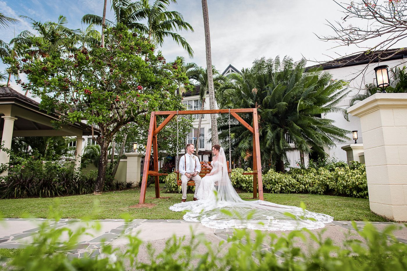 Garden wedding venue setup with white chairs and floral arch under palm trees.