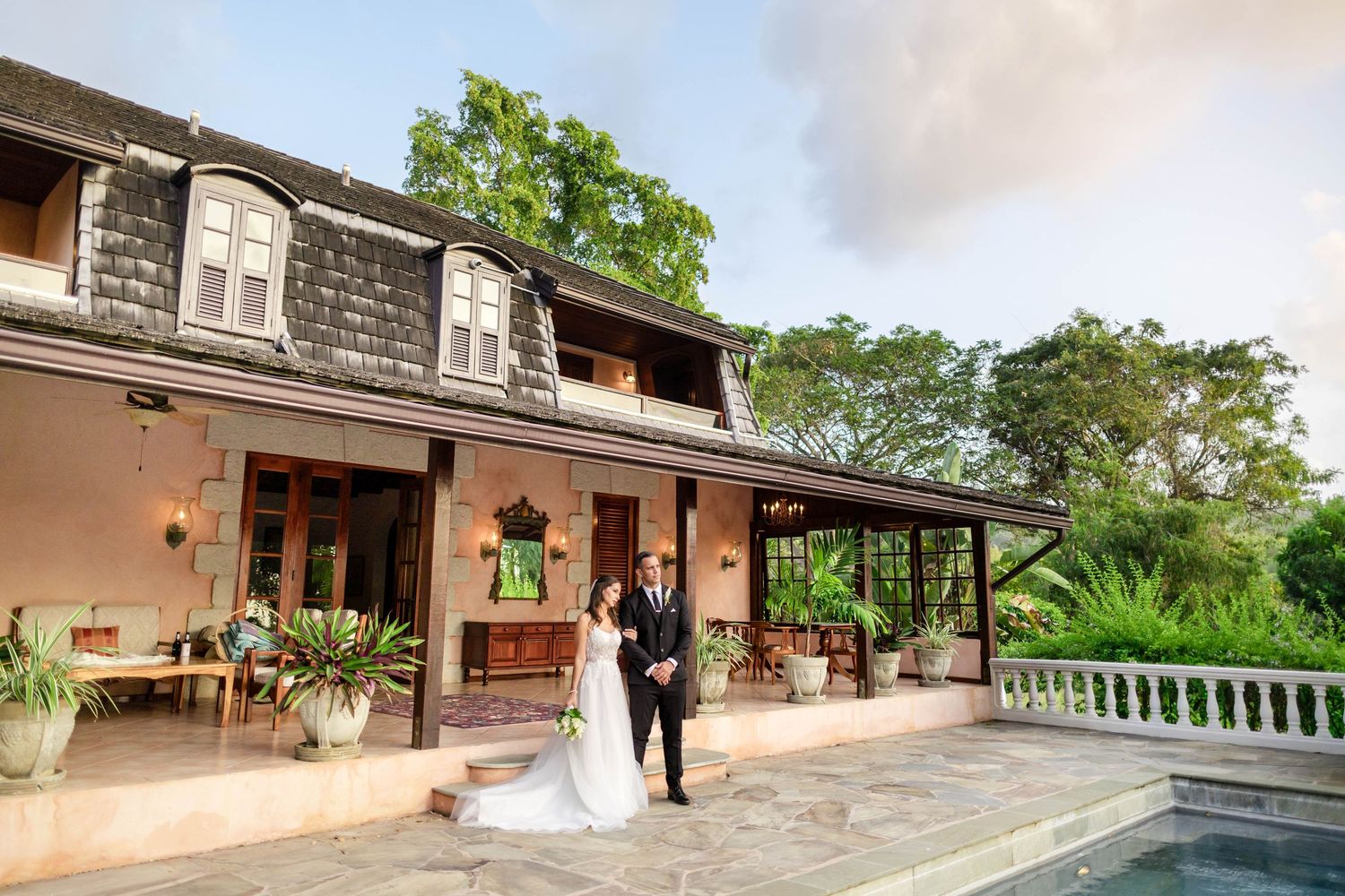 Bride and groom pose on a sunlit patio beside a pool at an elegant colonial-style wedding venue.