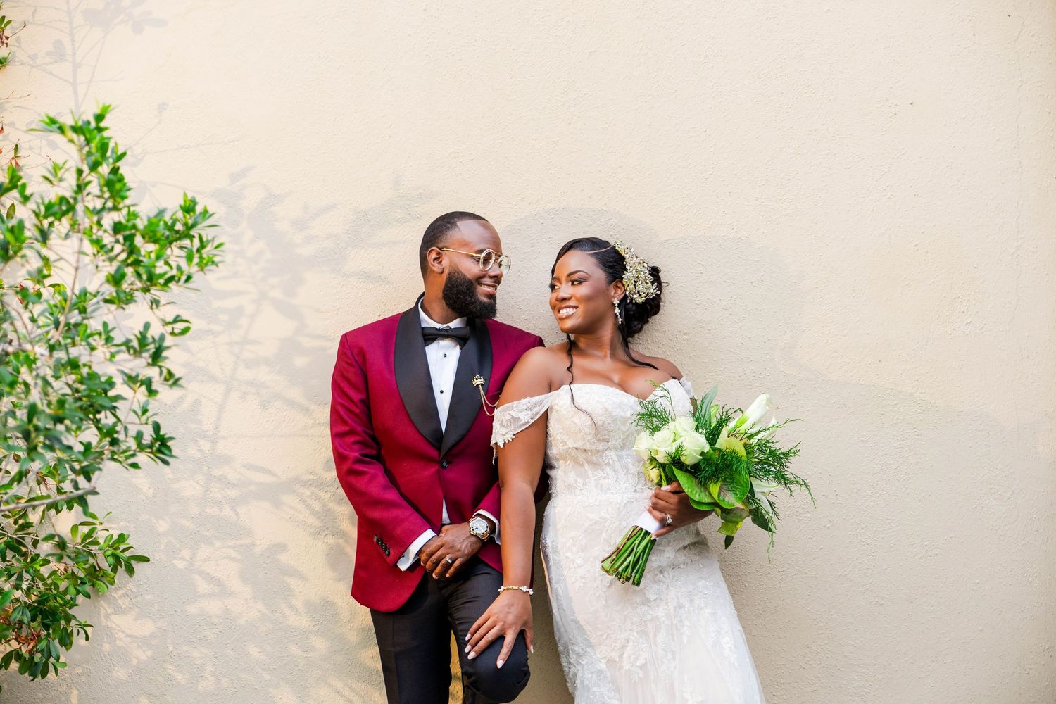 Smiling newlyweds pose against a beige wall, groom in burgundy tuxedo, bride in white off-shoulder gown holding bouquet.
