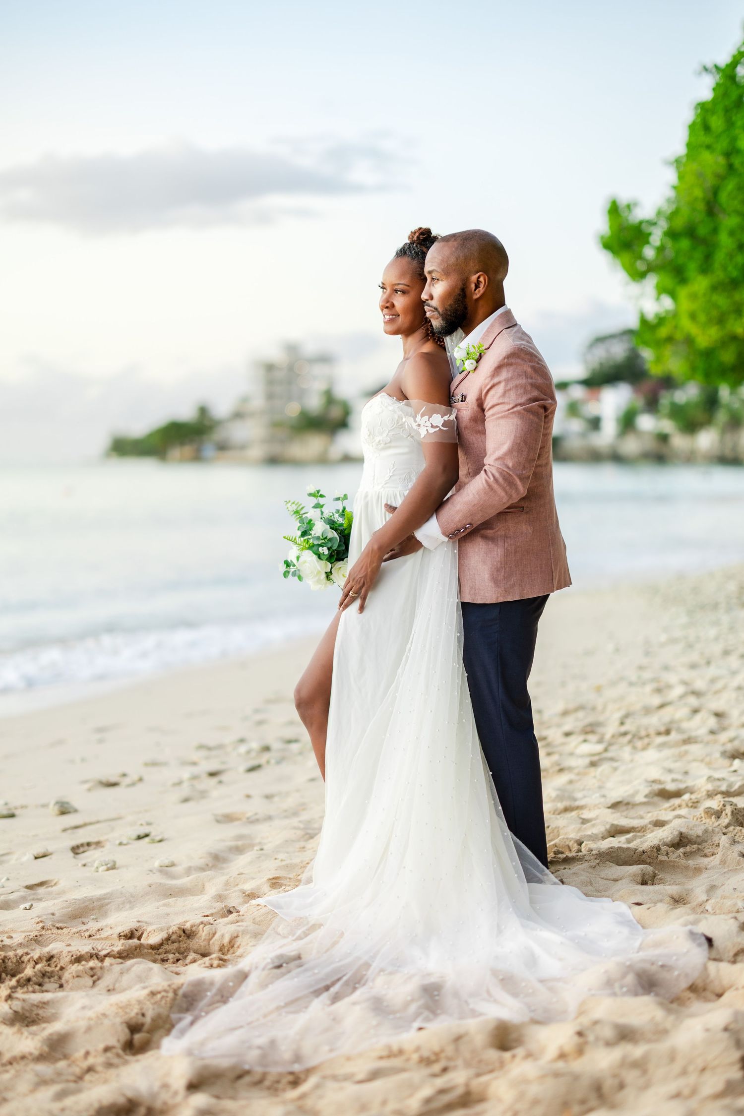 A bride and groom embrace on a sandy beach at sunset, she in a white gown holding flowers, he in a pink blazer.