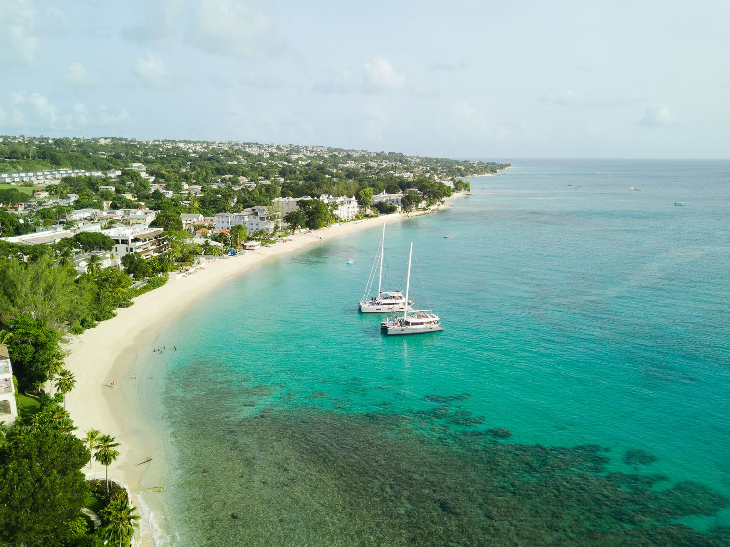 Aerial view of turquoise Caribbean waters with two sailboats anchored near a white sandy beach lined with tropical trees.