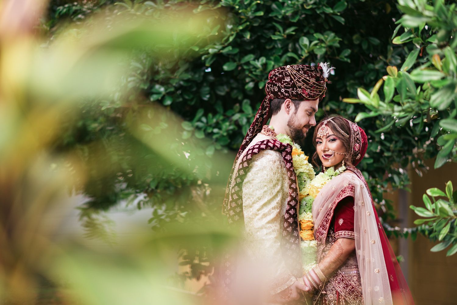 Indian bride and groom in traditional wedding attire share a romantic moment surrounded by lush greenery.