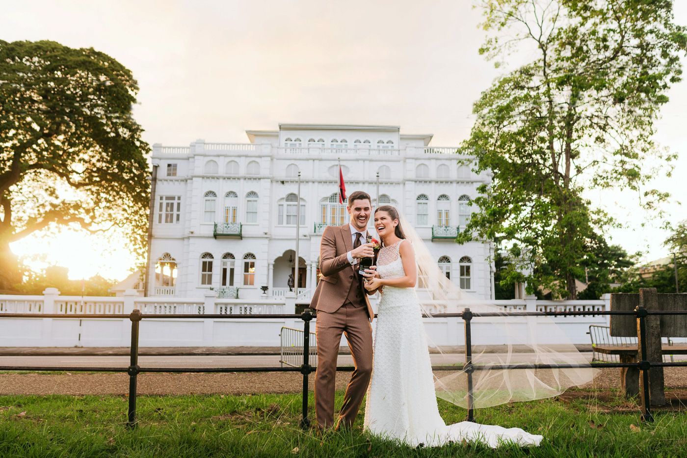 Bride and groom pose with baby in front of elegant white mansion at golden hour sunset.
