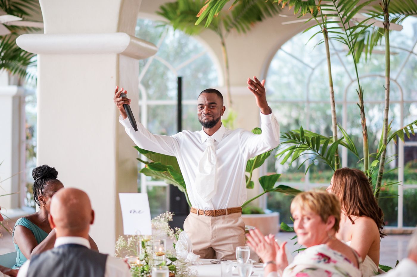 A guest delivers an enthusiastic speech during a wedding reception in an elegant venue with tropical decor.