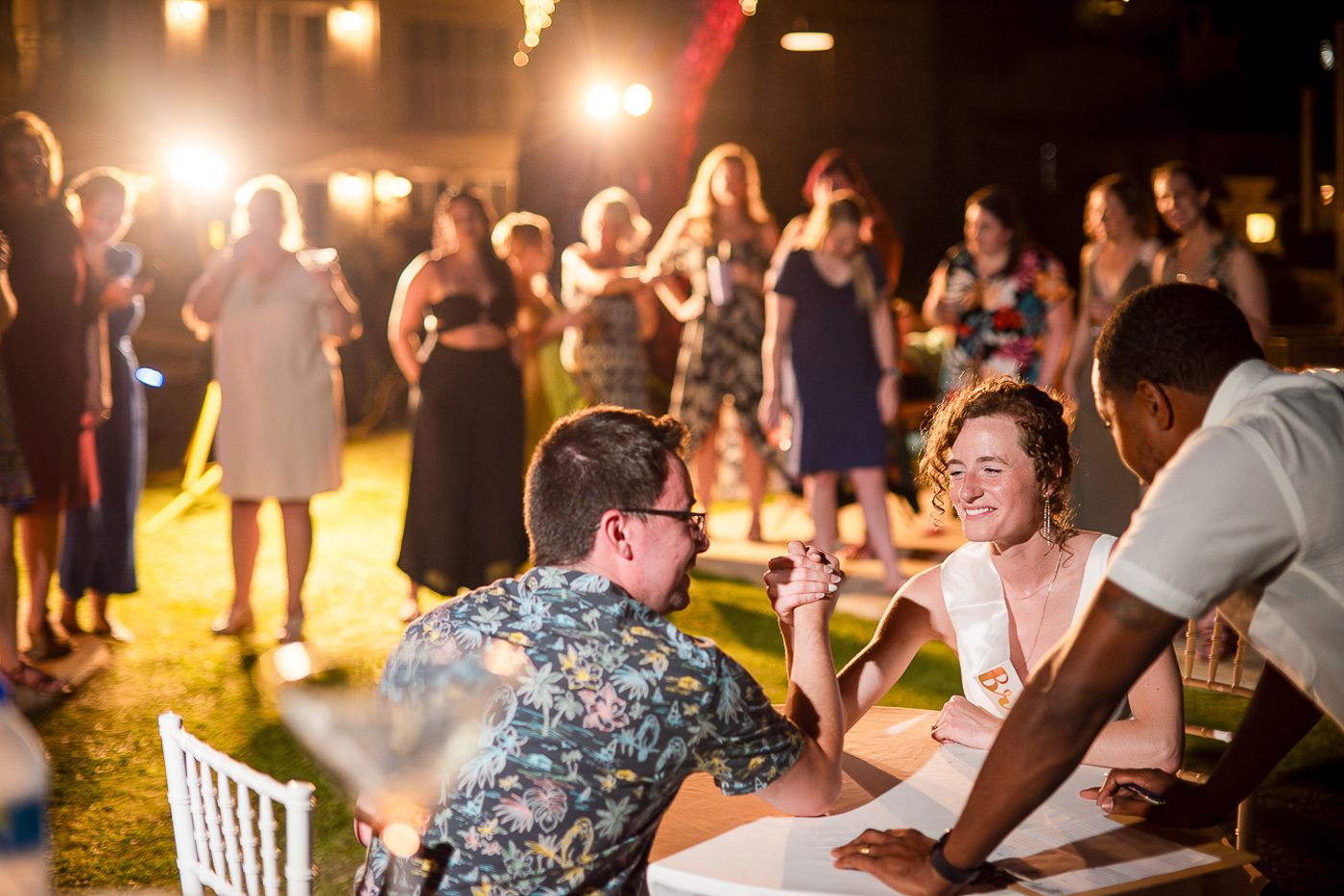 Outdoor evening party scene with people gathered around tables under ambient lighting.