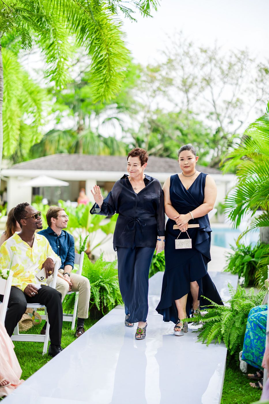 Wedding guests enjoy dynamic moments on a white walkway surrounded by tropical greenery.