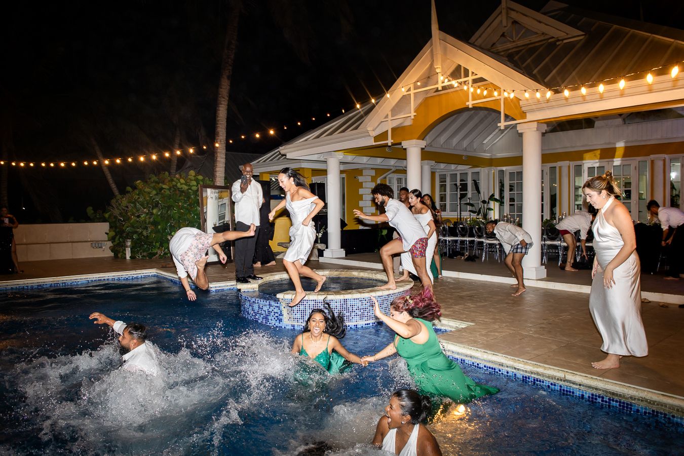 Wedding celebration with multiple guests splashing into pool at night near illuminated gazebo.