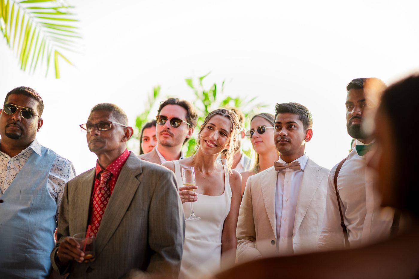 Wedding guests in light summer attire gathered outdoors under tropical foliage.