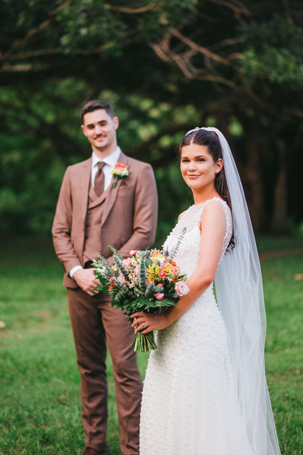 Wedding couple pose together outdoors in a garden setting with brown suit and white lace dress with floral bouquet.