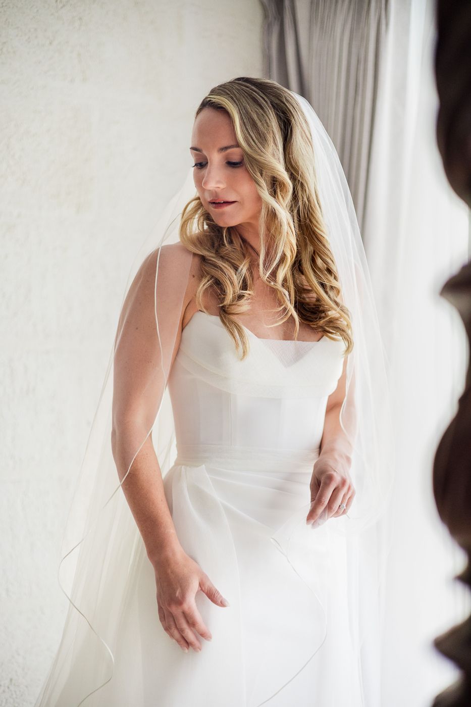 A bride in a strapless white wedding gown and veil gazes down gracefully near a bright window.