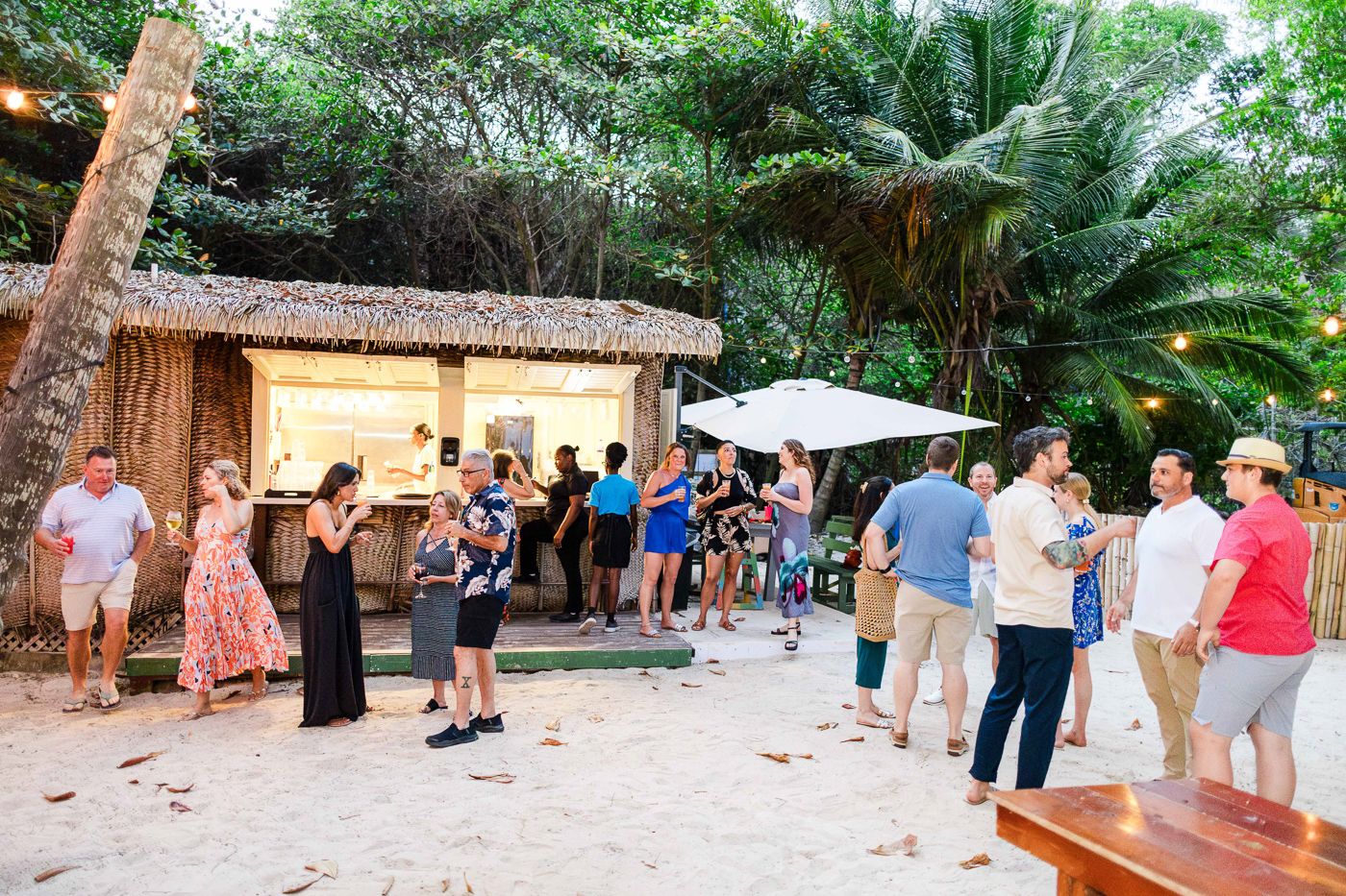 Guests mingle at a tropical beach bar with thatched roof, string lights, and lush greenery at an outdoor evening event.