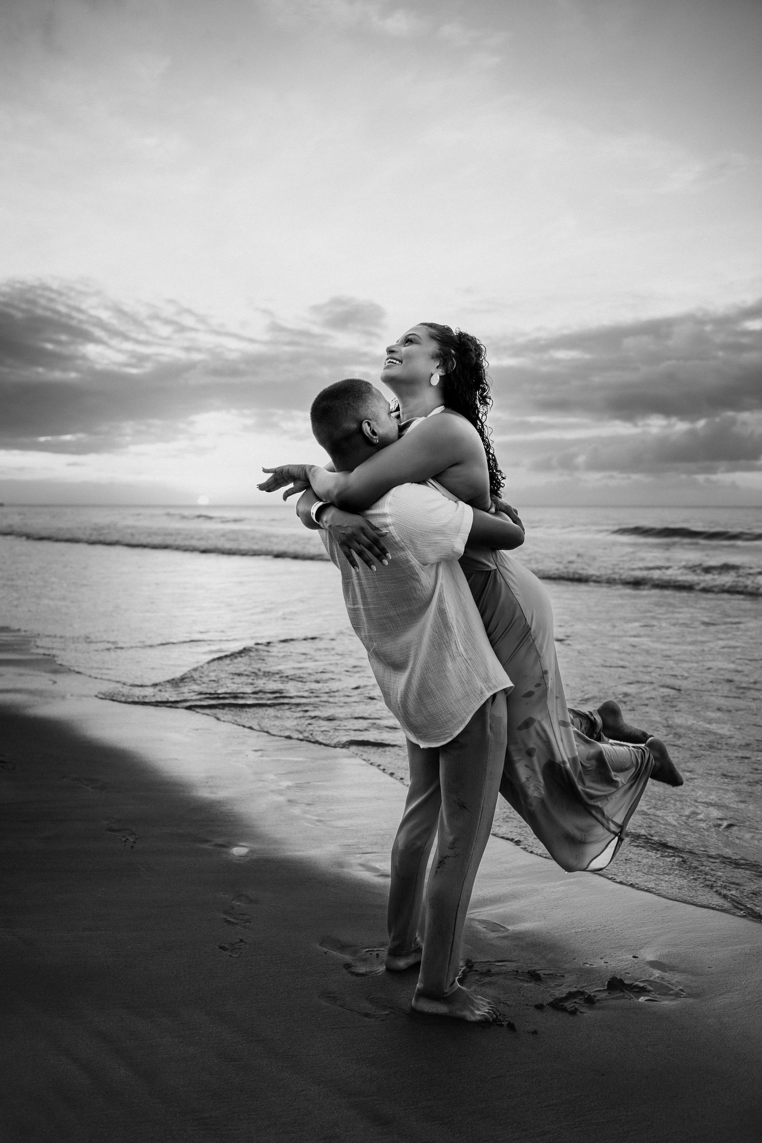 Black and white photo of a couple embracing joyfully on a beach at sunset, waves and cloudy sky in background.