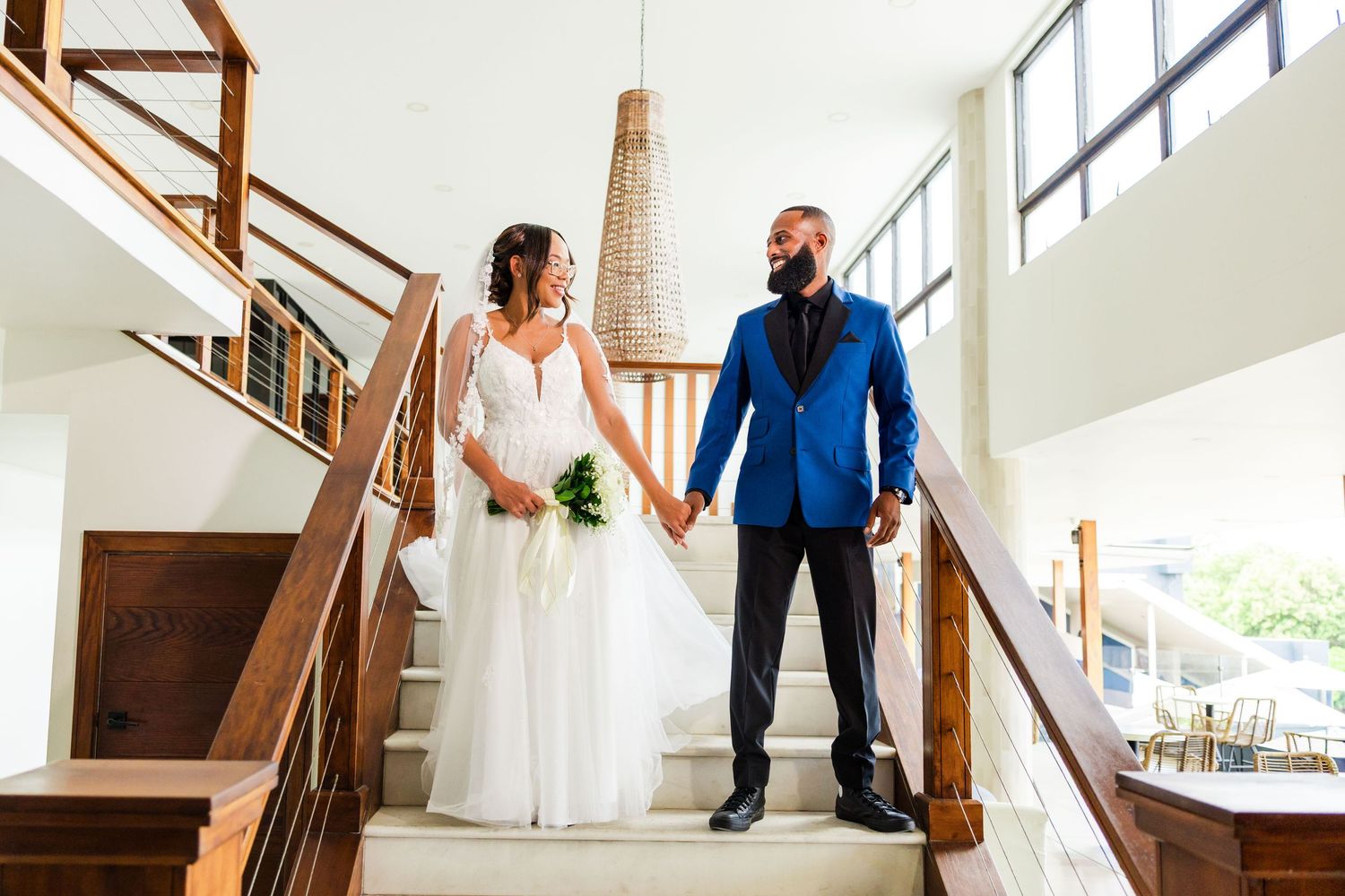 Bride and groom holding hands on staircase, bride in white gown with bouquet, groom in blue suit with black mask.