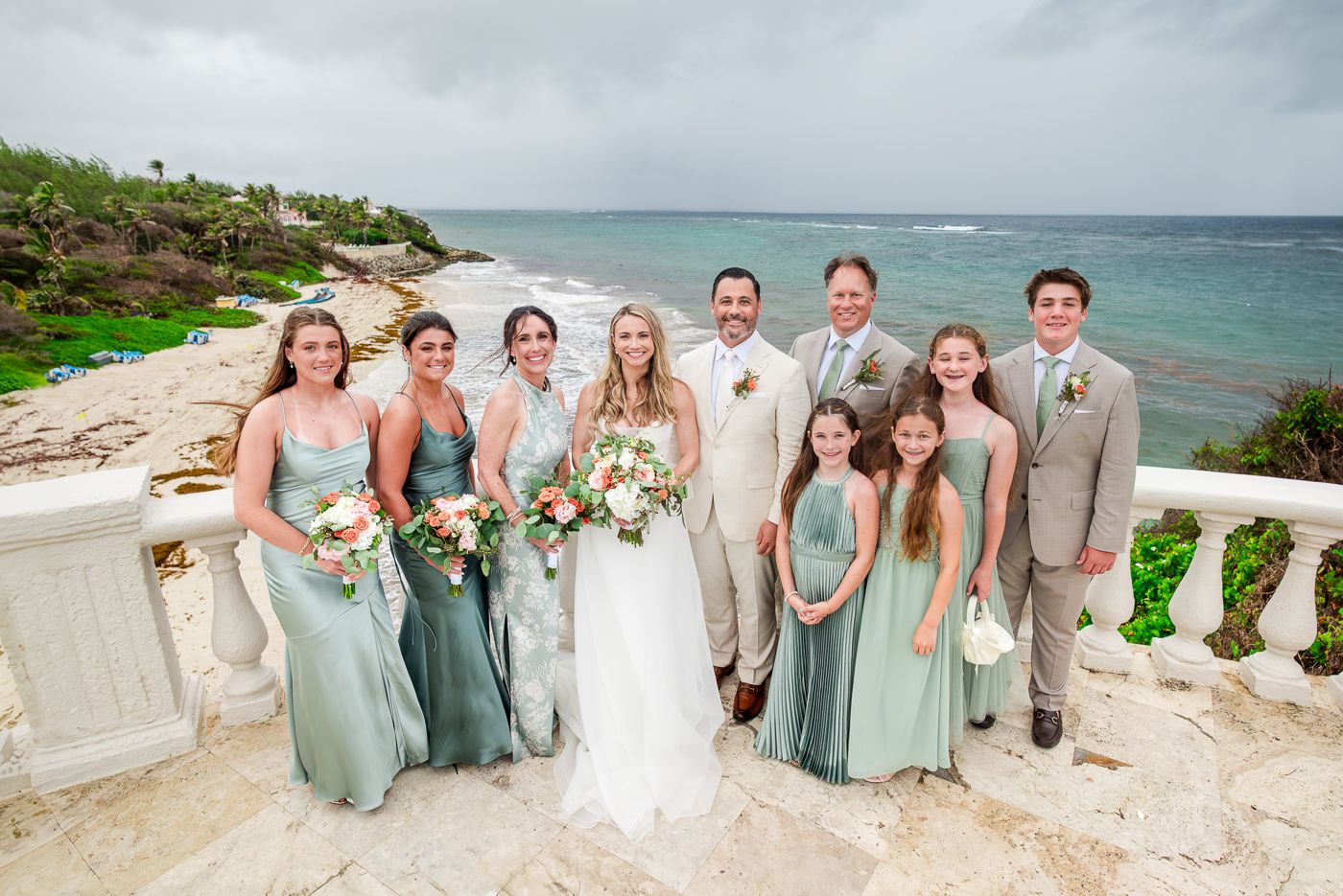 Wedding party posing on a beachfront terrace in the Bahamas, bridesmaids in sage green gowns, groomsmen in tan suits.