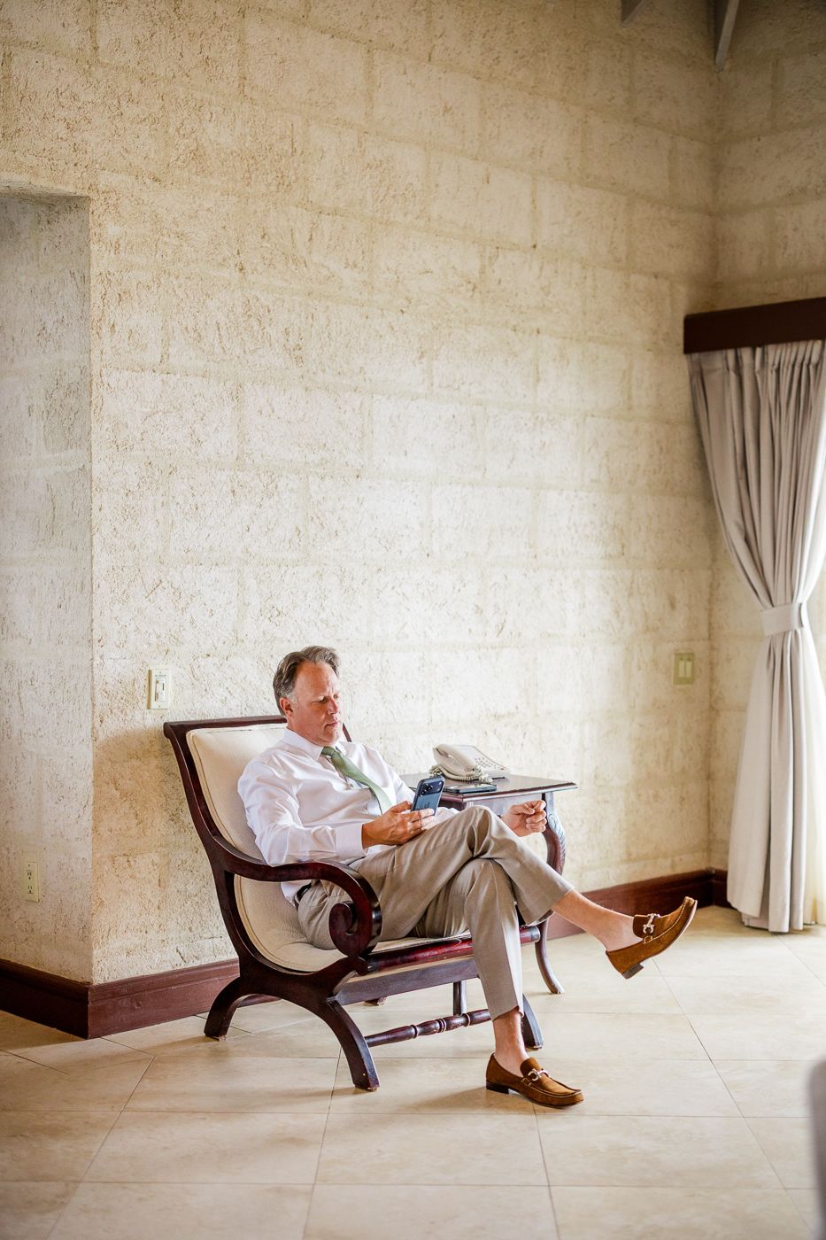 Businessman in white shirt and green tie relaxing in wooden armchair, reading in elegant stone-walled room.