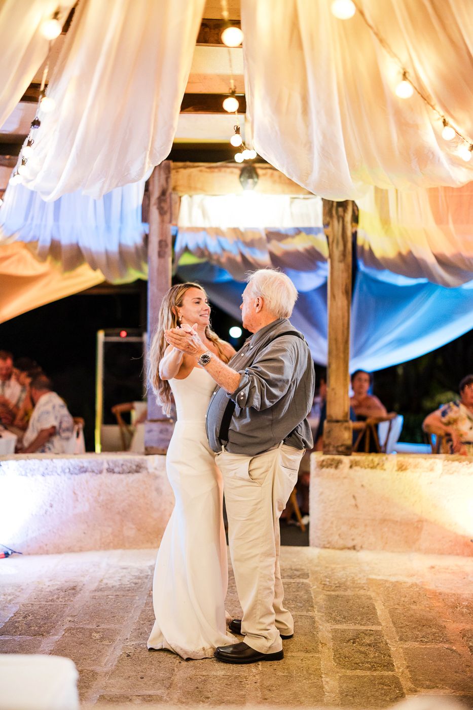 Bride in white gown shares father-daughter dance under string lights and draped fabric at outdoor wedding reception.