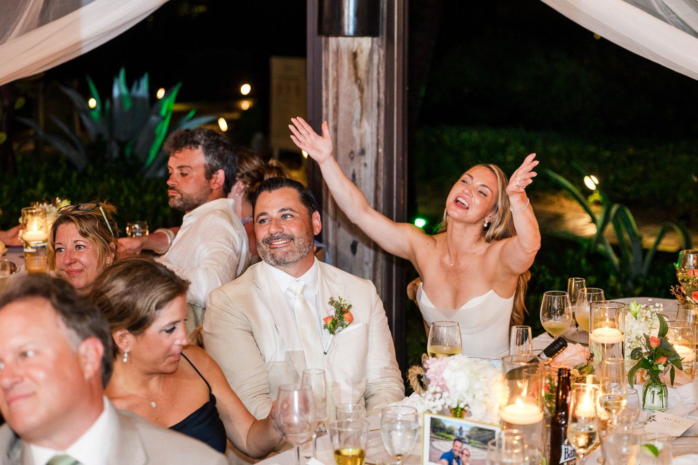 Bride laughing joyfully at wedding reception dinner table with guests celebrating under string lights.