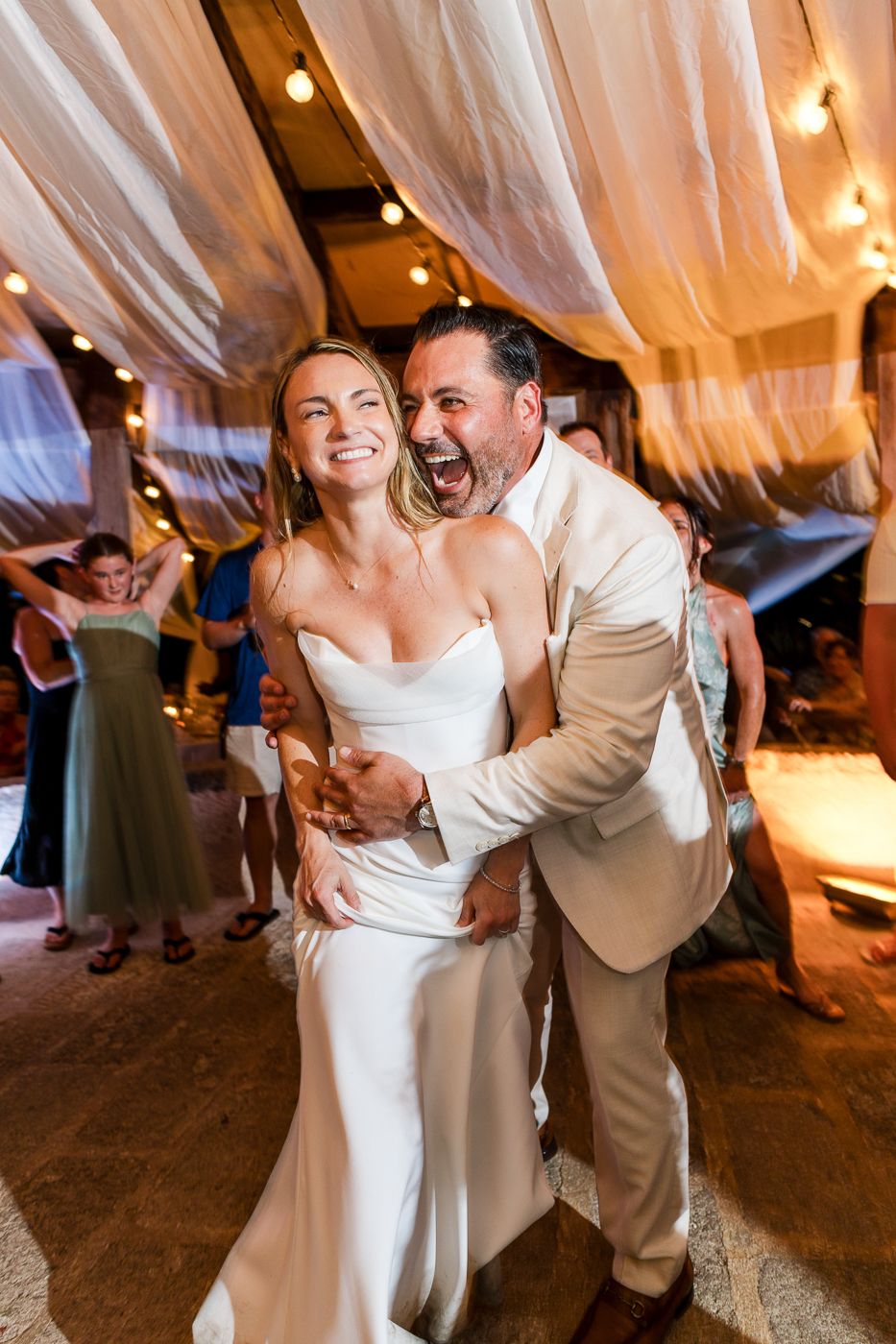 Joyful bride and groom dancing and laughing together at their wedding reception under warm string lights.