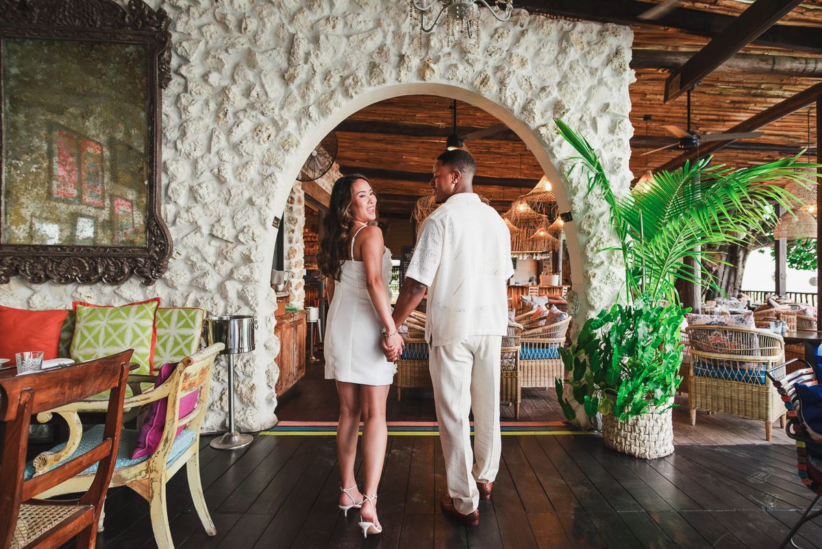 Couple in elegant attire walking through a coral stone archway in a tropical restaurant setting.