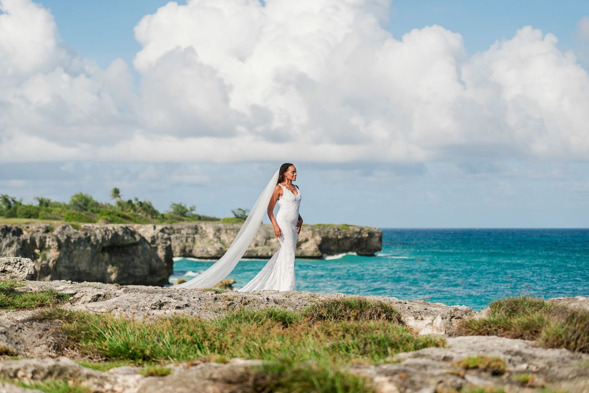 Bride in white gown and veil poses on rocky coastal cliffs with turquoise ocean and blue sky backdrop.