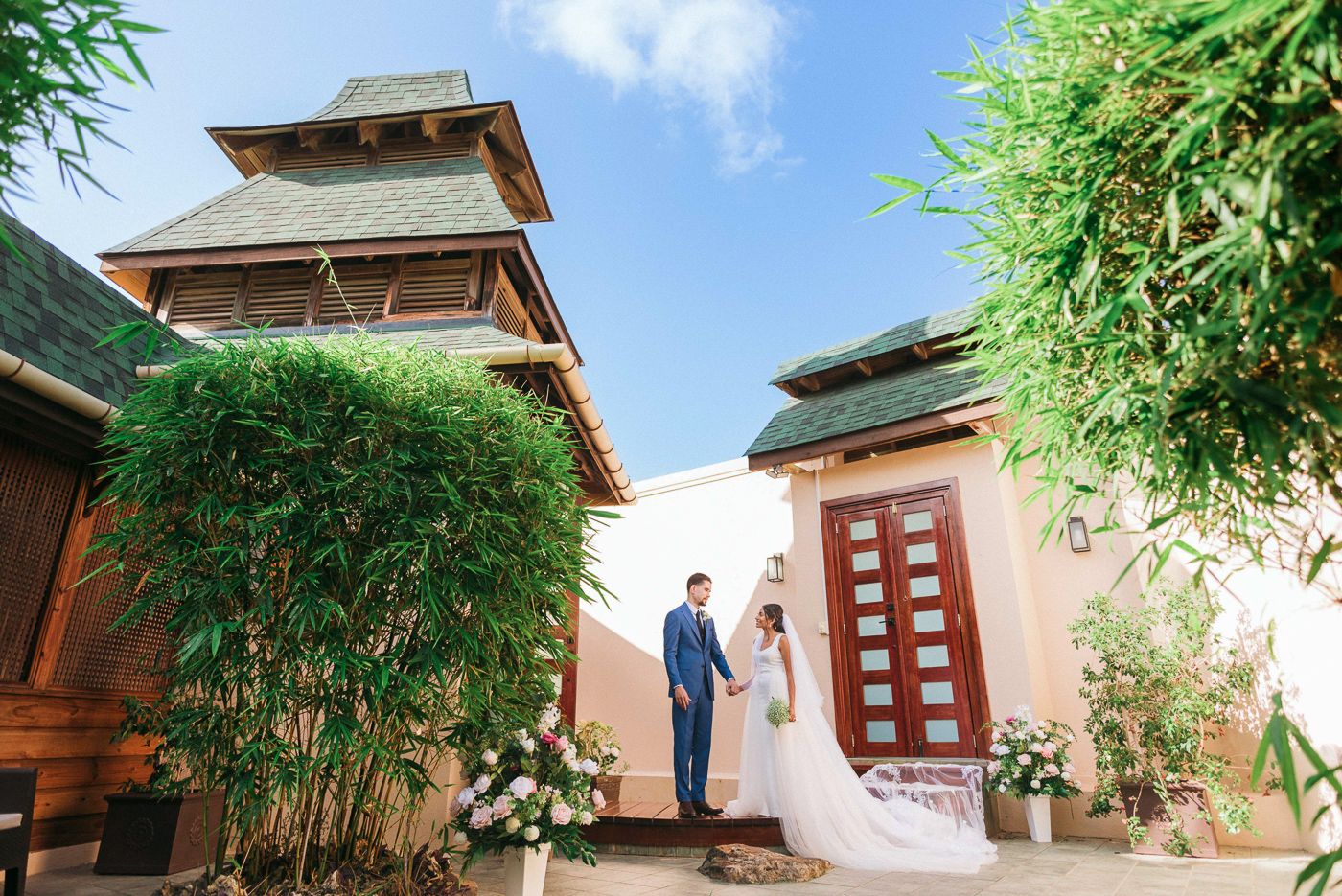 Wedding couple posing in front of elegant Thai-style villa with lush bamboo garden.