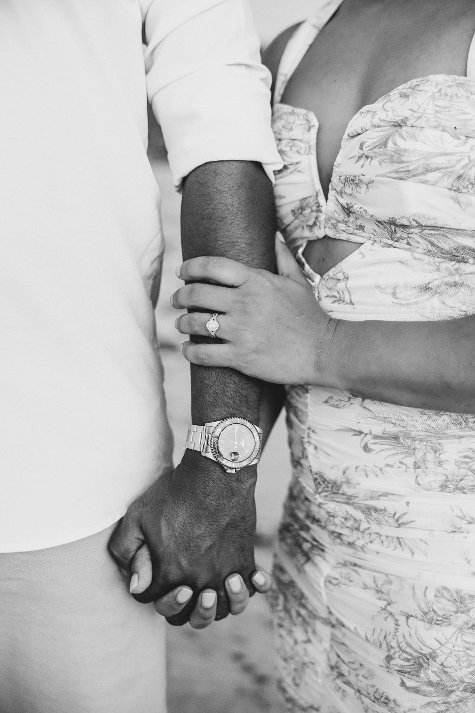 Black and white photo of a couple holding hands, woman wearing engagement ring and man wearing a watch.