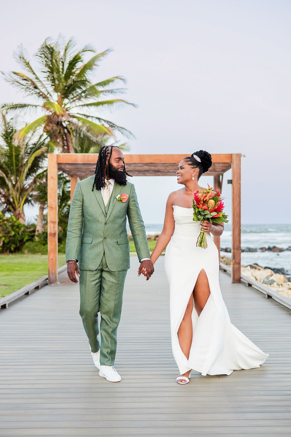 Bride and groom walk hand-in-hand on a beachside boardwalk, she in white gown, he in sage green suit.