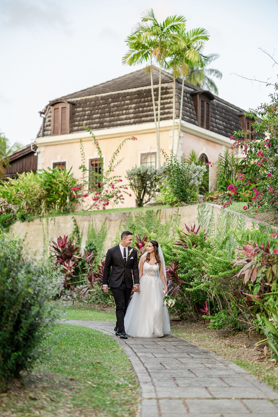 Bride and groom walking hand in hand along a stone path surrounded by lush tropical gardens near a pink villa.