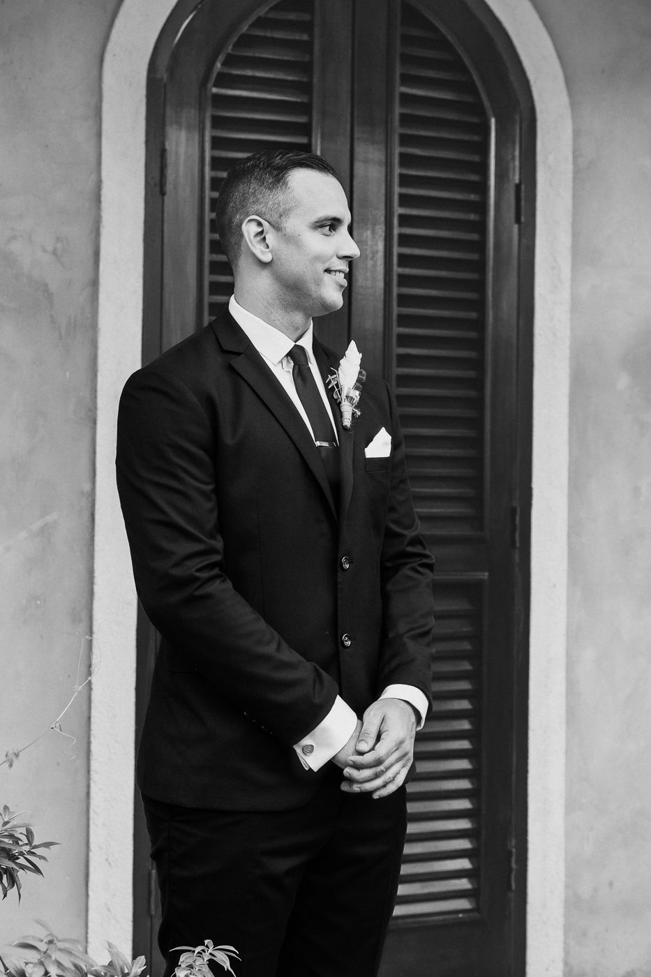 Dapper groom in black suit with boutonniere standing by arched shuttered door in black and white.