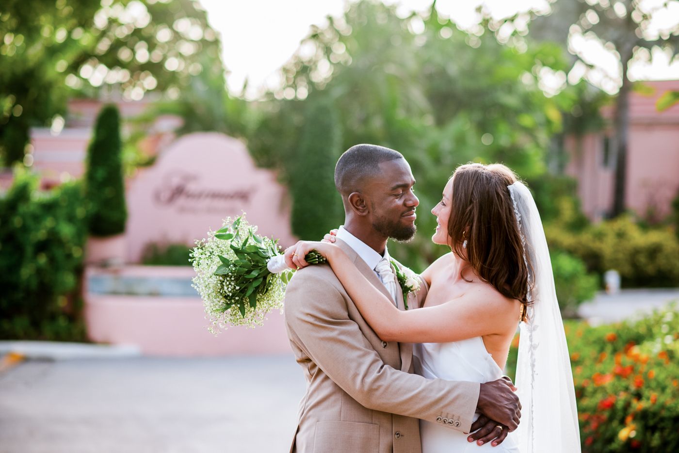 A couple embraces on a garden path surrounded by lush tropical landscaping and pink buildings.