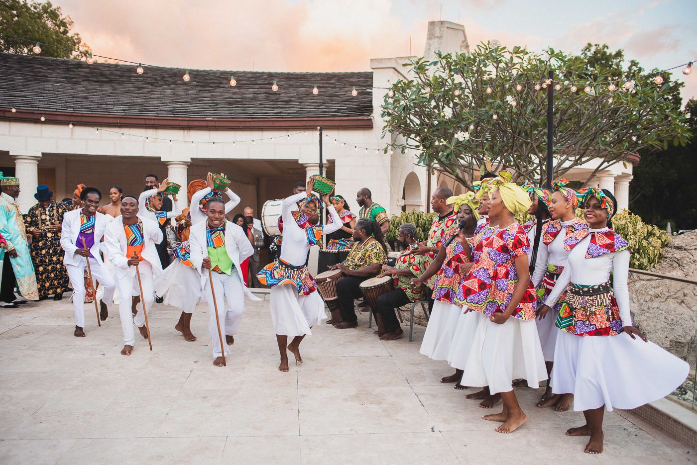 Wedding guests in white attire dancing and celebrating on a beach during sunset.