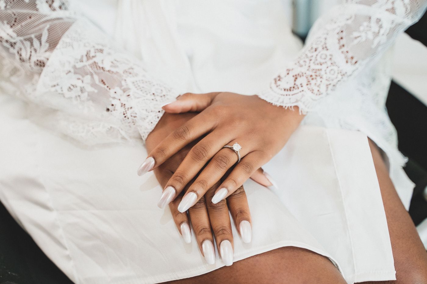 Close-up of folded hands with manicured nails wearing engagement ring on white lace dress.