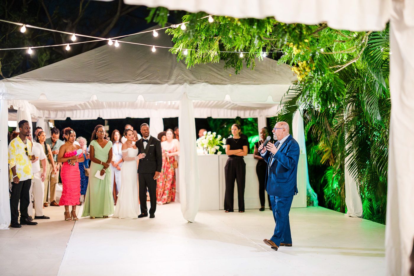 Wide view of a white tent wedding reception setup with string lights and tropical foliage.